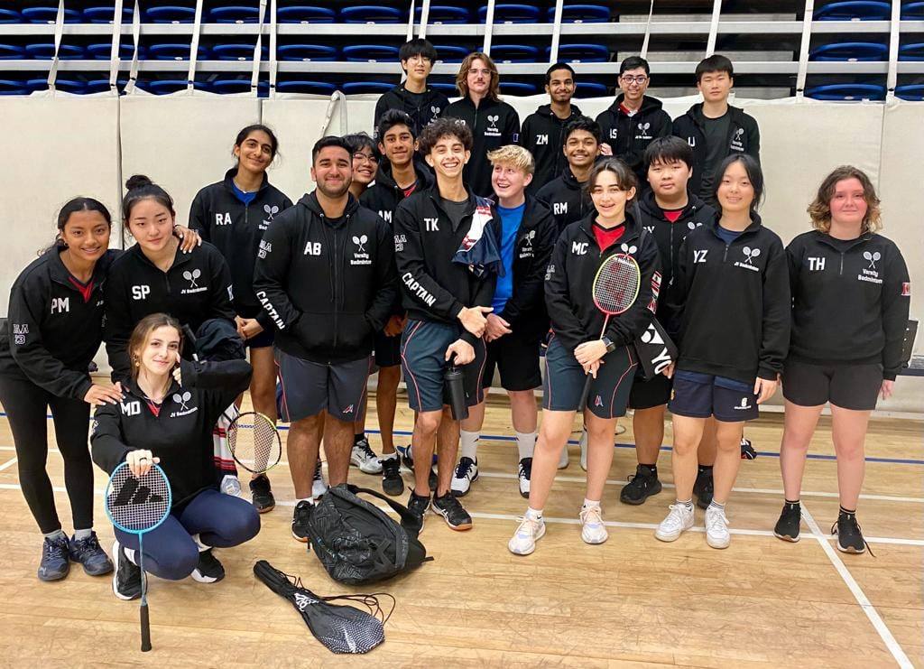 A badminton team in matching black hoodies posing in a sports hall.