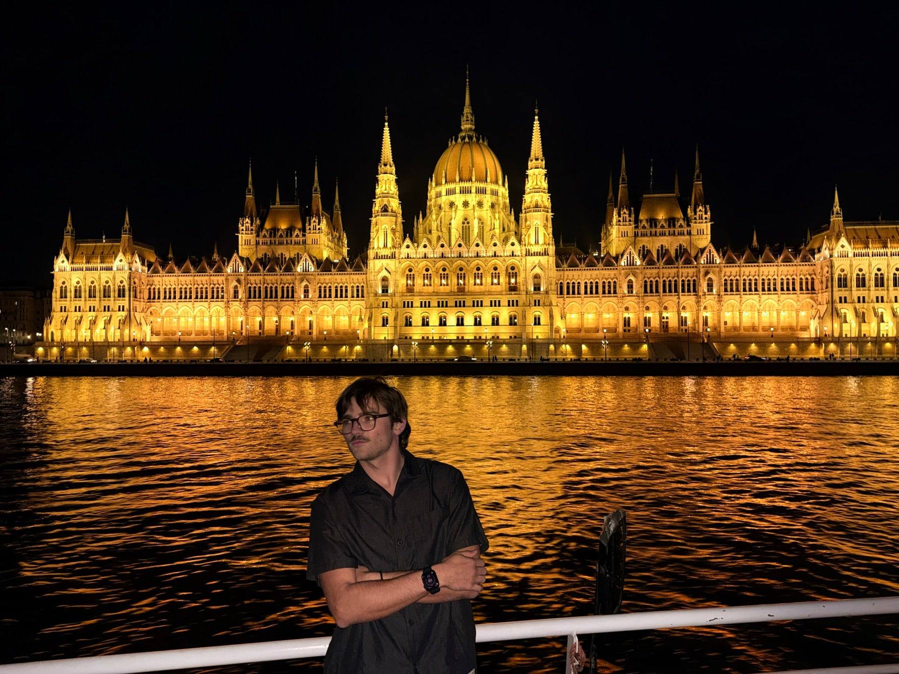 Sinclair on a river boat at night, arms crossed, in front of the illuminated Hungarian Parliament in Budapest.