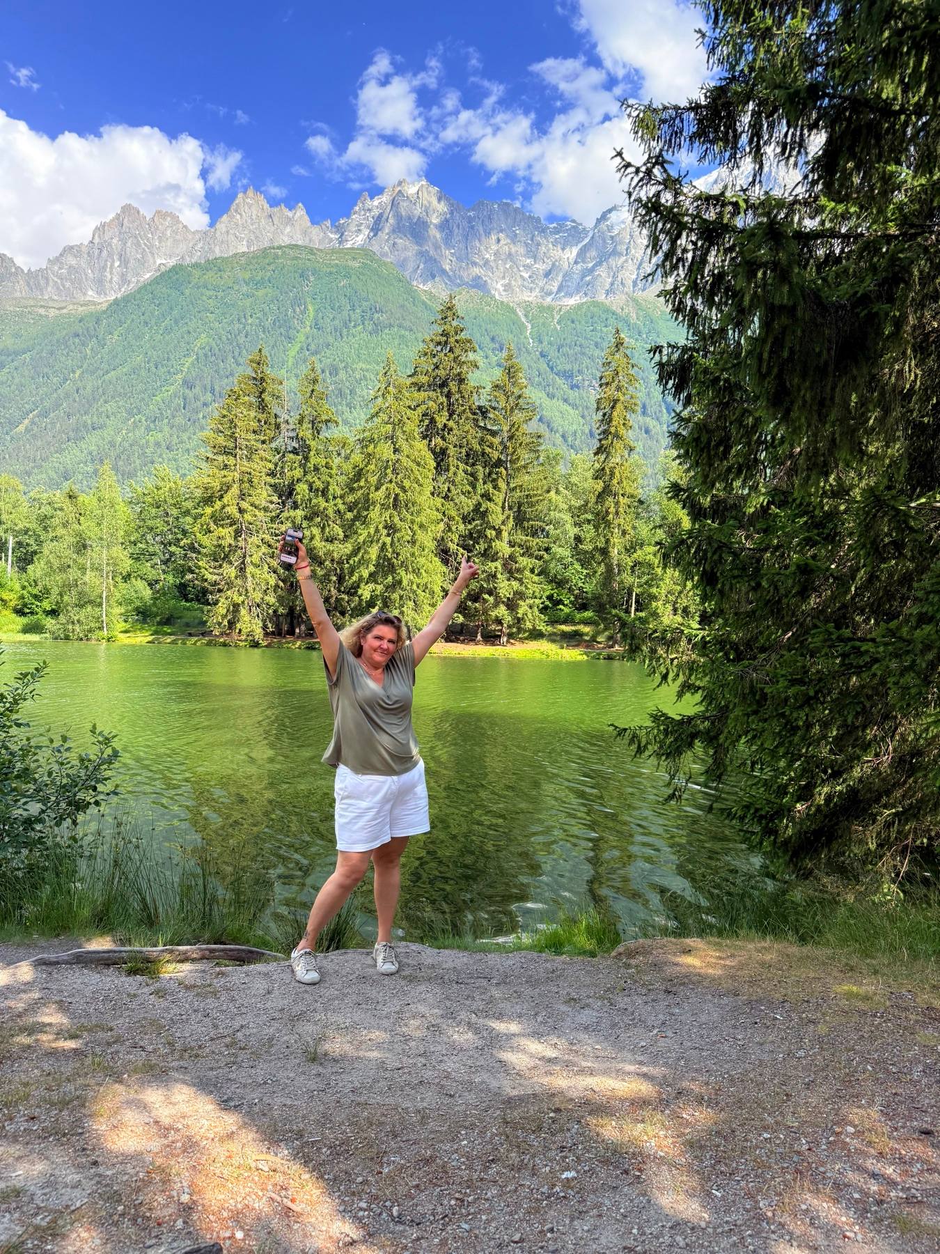Carine smiling with her arms raised beside a lake in the Alps.