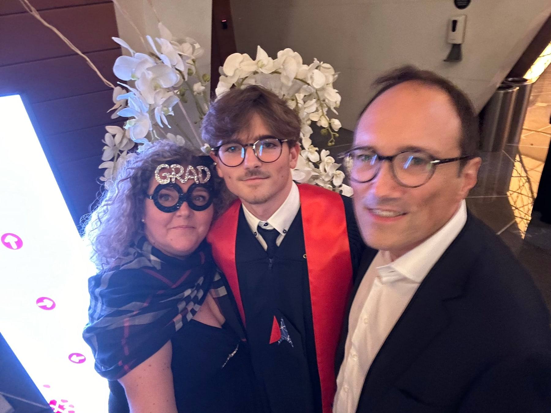 Sinclair in a red graduation stole with two family members, one wearing GRAD novelty glasses.