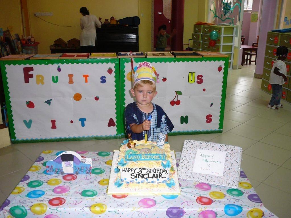 Sinclair at about three, in a paper birthday crown, beside a cake reading “Happy 3rd Birthday Sinclair” in a bright classroom.