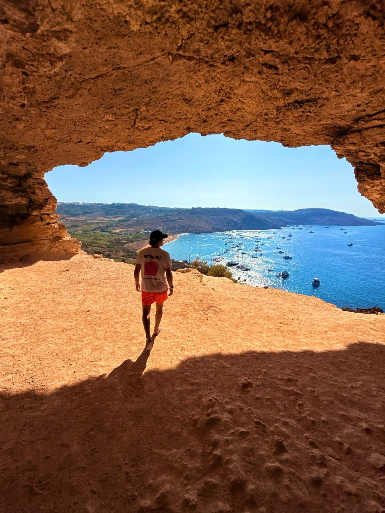 Looking out from a sandstone cave to a turquoise bay in Malta, Sinclair walking towards the light.