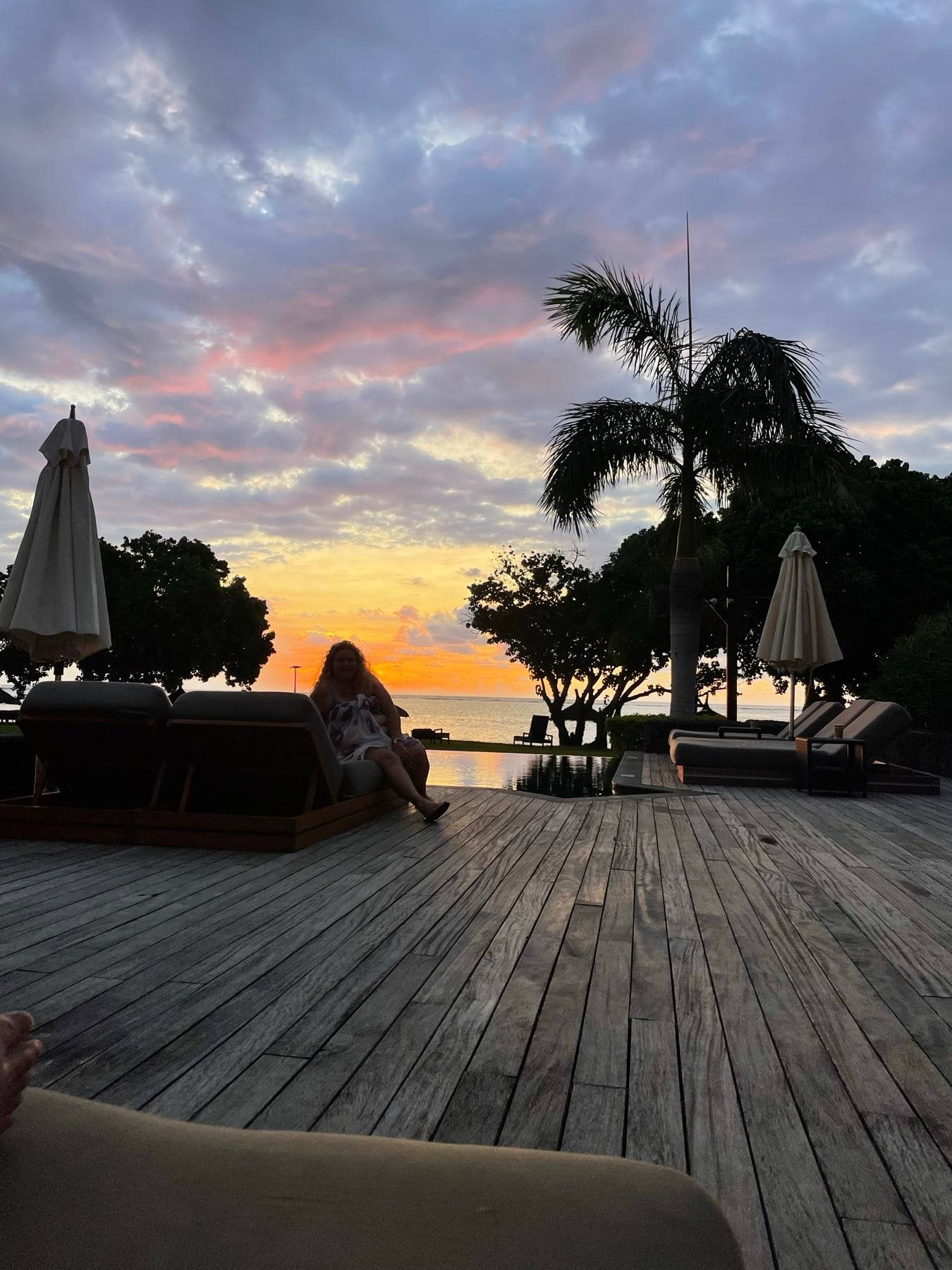 A wooden deck with palms and umbrellas silhouetted against a sunset.