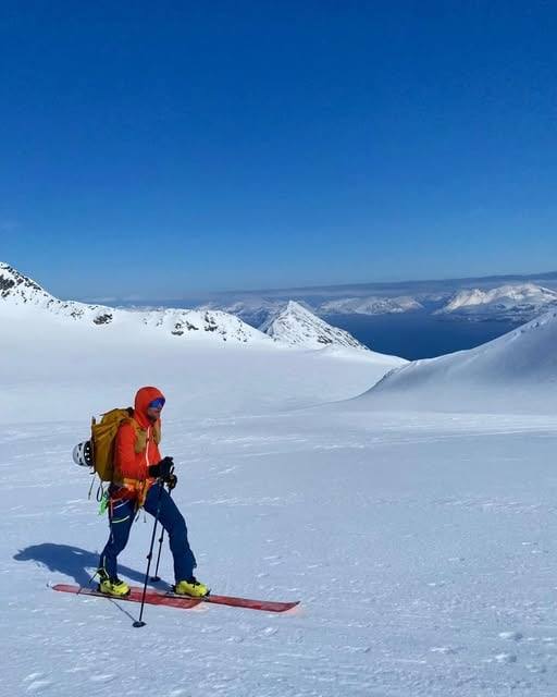 A ski tourer in a red jacket crossing a wide glacier under blue sky.
