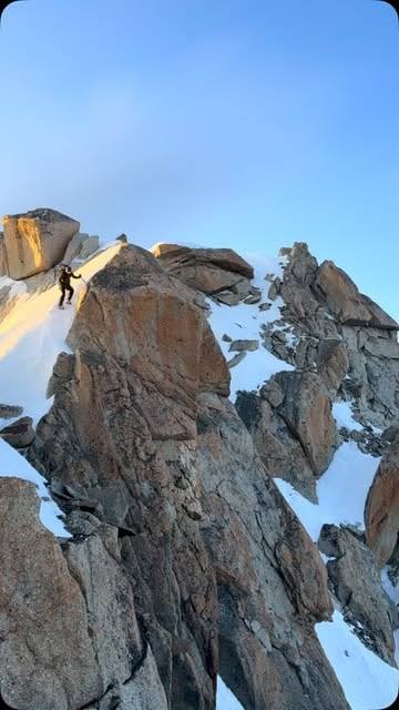 A climber high on a sunlit granite ridge above snow.