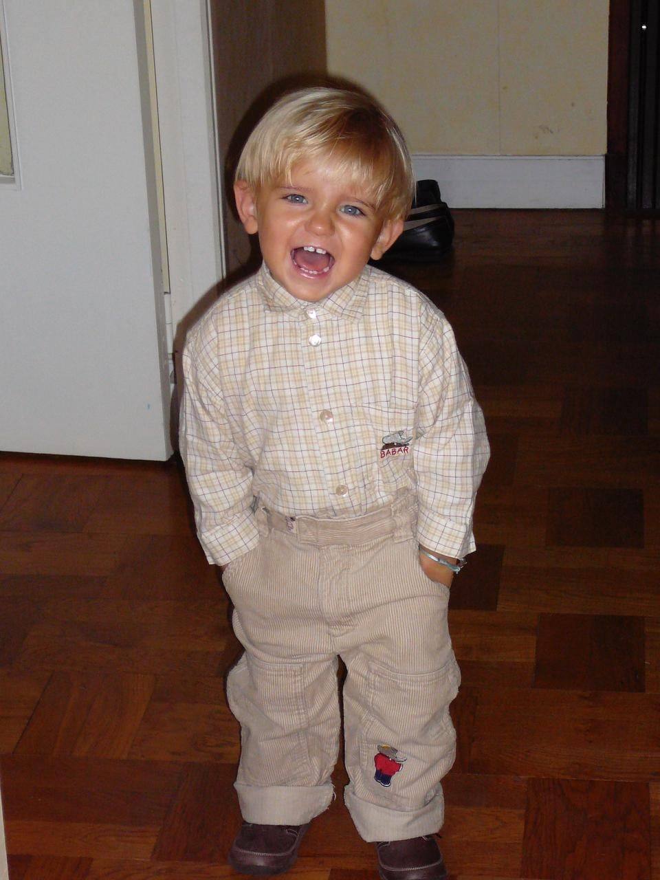 Sinclair as a laughing blond toddler in a checked shirt and cord trousers, hands in pockets, on a parquet floor.
